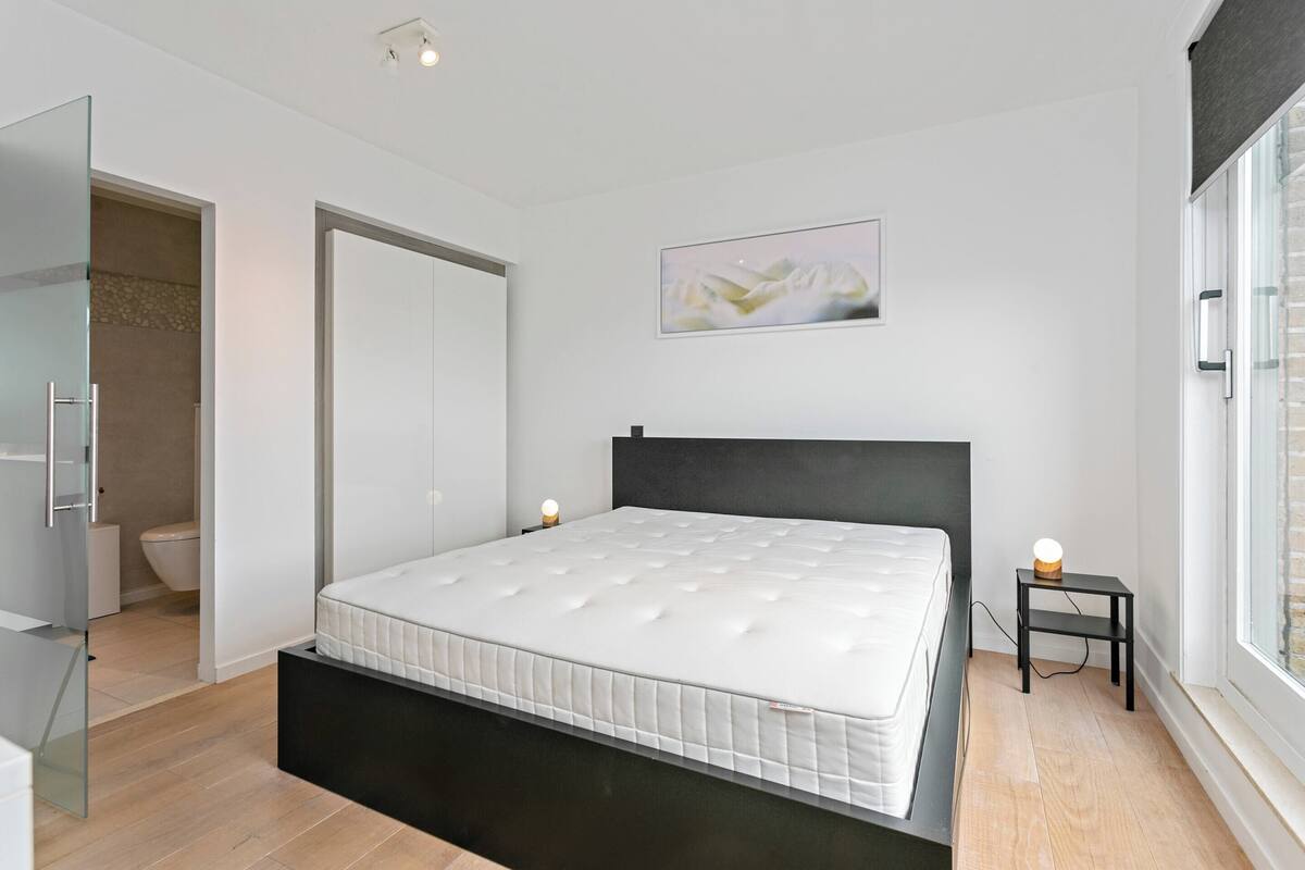 A modern bedroom with a bed, a bedside table with a lamp, a painting on the wall, and a window.
