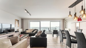 A modern living room with a sofa, a coffee table, a TV, and a dining area with chairs and a table. - Penthouse in Blankenberge Near the Beach (Blankenberge)