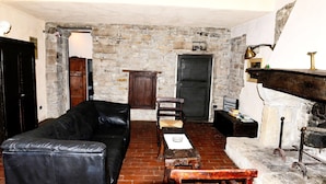 A room with a stone wall, a fireplace, a black leather sofa, a wooden chair, and a small table with books. - Farmhouse in Modigliana With Scenic Views (Modigliana)
