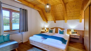 A bedroom with a wooden ceiling, a bed with a blue blanket, a nightstand with a lamp, and a window with curtains. - Vacation apartment Sonnenblume, up to 2 persons, top floor, 50 sqm (Waging am See)
