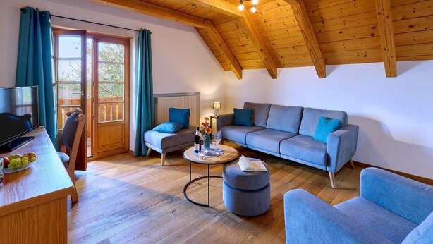 A modern living room with a wooden ceiling, a grey sofa, a blue armchair, a wooden dining table with fruit, and a TV. - Vacation apartment Schlüsselblume, up to 4 persons, attic, 65 sqm (Waging am See)