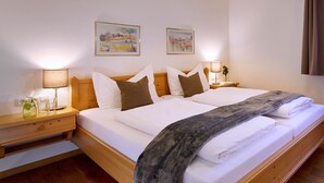 Ein Doppelbett mit weißen Leinentüchern und einer grauen Tagesdecke, ein hölzernes Kopfende, Nachttische mit Lampen sowie zwei gerahmte Kunstwerke an der Wand.