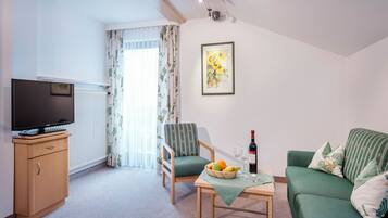 Een woonkamer met een televisie, een fauteuil, een bank, een tafel met fruit en wijn, en een schilderij aan de muur.