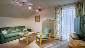 Una sala de estar con un sofá verde, una silla de madera, una mesita, un radiador, una ventana con cortinas y un televisor.