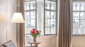 Ein Zimmer mit einer Ledersofa, einem runden Holztisch, einer Lampe, einer Vase mit Blumen und großen Fenstern mit Vorhängen.