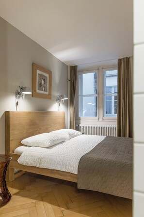A bedroom with a wooden headboard, a bed with white bedding, a bedside table, a framed picture on the wall, and a window with curtains. - Bijou in the middle of the old town of Bern (Bern)