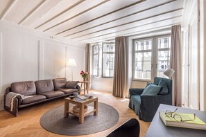 A living room with a sofa, a coffee table, and a chair. There is a window with curtains, a lamp, and a view of buildings outside. - Bijou in the middle of the old town of Bern (Bern)