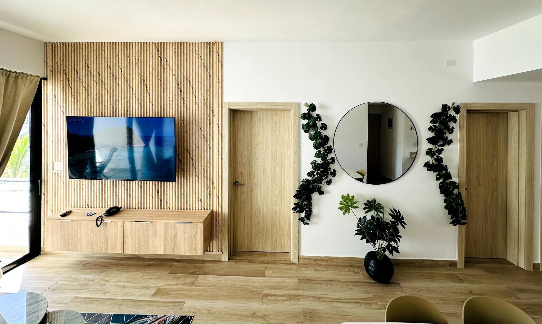 En moderne stue med et tv monteret på en væg af træpaneler, et rundt spejl og en potteplante.