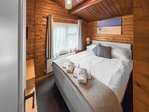 Ein schlicht eingerichtetes Schlafzimmer im Chalet-Stil mit Bett, Kissen, einem Nachttisch, einer Lampe, einem Fenster mit Vorhängen und einem Bild an der Wand.