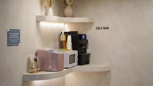 Ein Selbstbedienungsbereich mit einer Mikrowelle, einer Kaffeemaschine und einer Vase mit getrockneten Blumen auf einer weißen Theke.