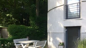 Une terrasse avec une table et des bancs, entourée de verdure et bordée d’un bâtiment doté d’une porte et d’une fenêtre.