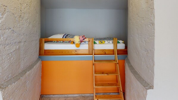 Una cama individual con escalera de madera, un colchón y una almohada con un personaje de dibujos animados, y una pared de textura rugosa.