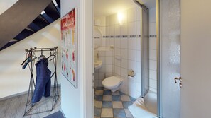 Ein Badezimmer mit Toilette, Waschbecken und Dusche. An der Wand hängt ein Poster, und ein Schuhregal steht bereit.