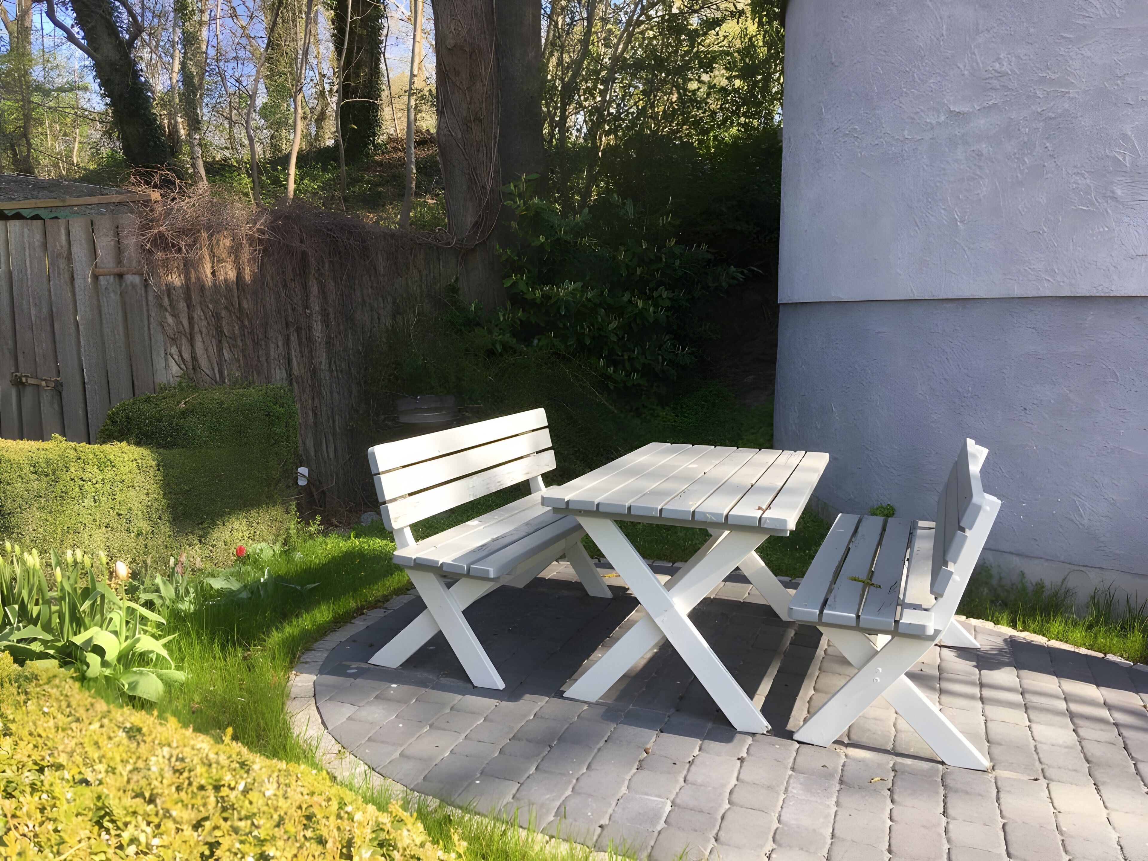 Zona d'seients exterior blanca amb taula i bancs, envoltada d'un pati pavimentat i vegetació.