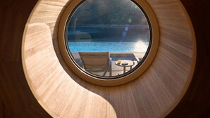Una ventana circular con vista a una masa de agua y una pequeña mesa con una silla.