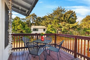 A wooden deck with a metal table and chairs, a potted plant, and a view of a house and trees. - Movie Room + Fire Pit + Deck with Views + Walk to the Beach | Magic Tree House (Manzanita)