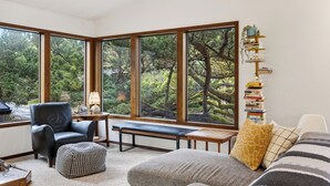 A cozy room with a large window, a bench, a chair, a small table, and a bookshelf. - Movie Room + Fire Pit + Deck with Views + Walk to the Beach | Magic Tree House (Manzanita)