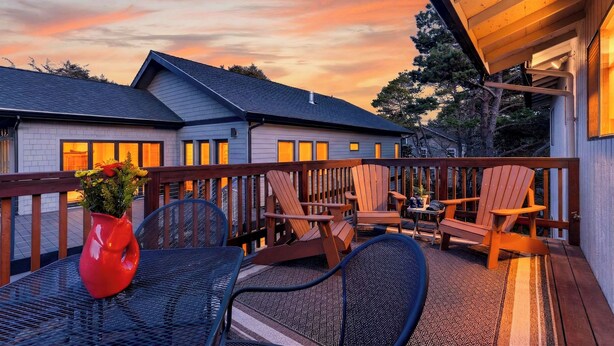 A wooden deck with outdoor furniture, a red vase with flowers, and a house in the background. - Movie Room + Fire Pit + Deck with Views + Walk to the Beach | Magic Tree House (Manzanita)