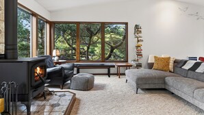 A modern living room with a fireplace, large windows, a sofa, a bench, and a bookshelf. - Movie Room + Fire Pit + Deck with Views + Walk to the Beach | Magic Tree House (Manzanita)