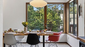 A dining area with a table set for breakfast, a large window with a view of trees, and a hanging light fixture. - Movie Room + Fire Pit + Deck with Views + Walk to the Beach | Magic Tree House (Manzanita)