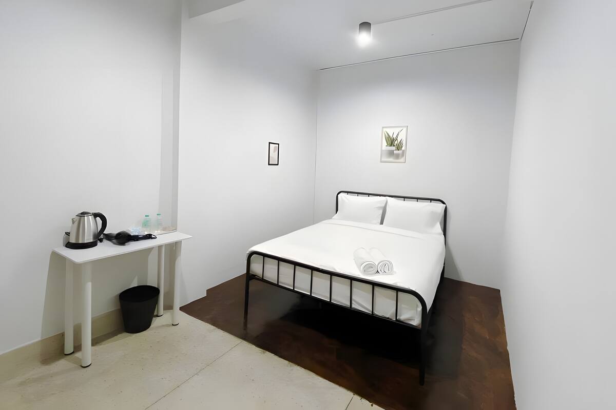 Ein Schlafzimmer mit einem Bett, einem kleinen Schreibtisch, einem Mülleimer und einem an der Wand befestigten Regal mit einer Pflanze.