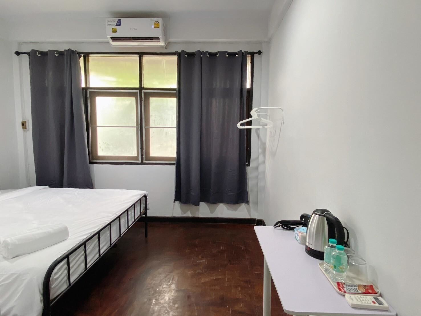 A room with a bed, an air conditioning unit, a window with curtains, a table with a kettle, water bottles, and a small tray with snacks.