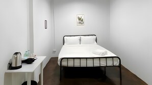 Minimalistinen hotellihuone, jossa on yksi sänky, pieni yöpöytä, vedenkeitin ja seinällä oleva ruukkukasvi.