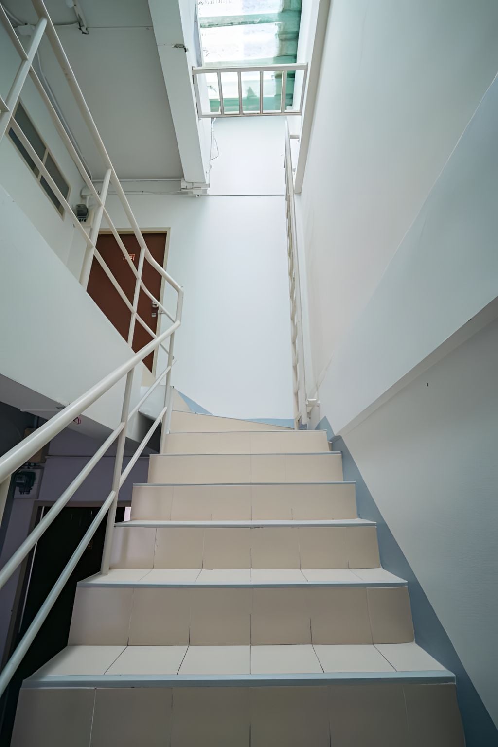 Uma escadaria com degraus brancos e corrimão de metal, que leva a um patamar com janela.