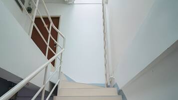 Uma escadaria com degraus brancos e corrimão de metal, que leva a um patamar com janela.