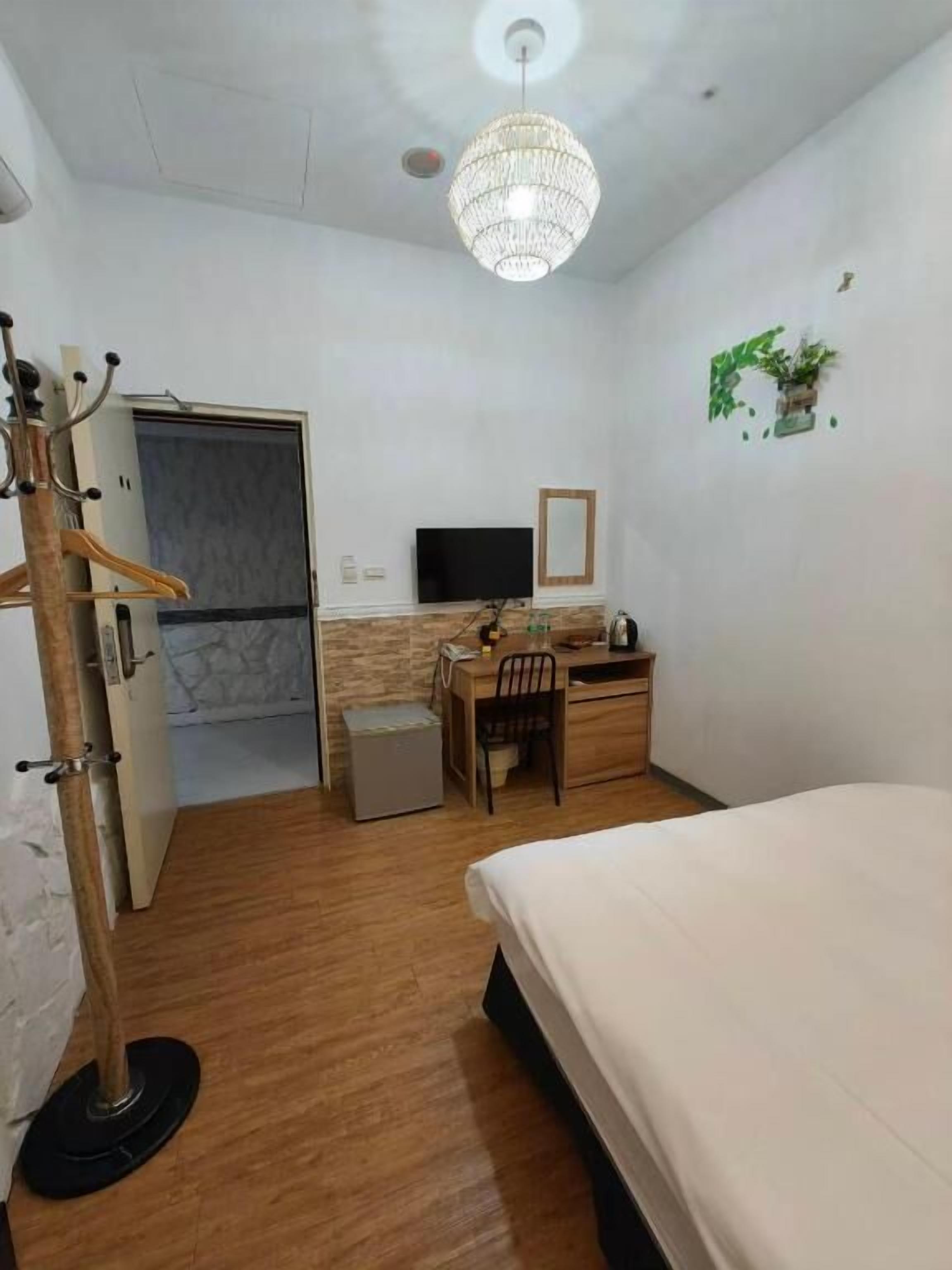 Una habitación de hotel con una cama, un escritorio con silla, televisión, un cubo de basura pequeño y un perchero.