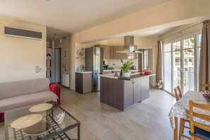 A modern kitchen with a central island, a dining area with a table and chairs, a living room with a sofa, and large windows providing natural light. - Irodotou Two Storey Apartment Magic View (Kefalonia)