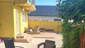 Un patio al aire libre con mobiliario de mimbre, un edificio amarillo y una vista de un edificio con tejado de baldosas al fondo.