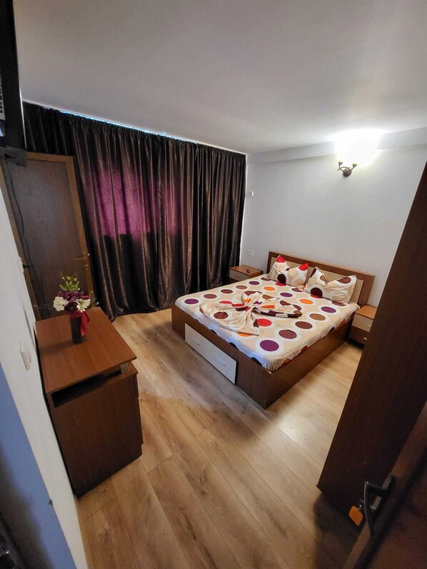 A bedroom with a bed, a wooden nightstand, a desk, a chair, a wardrobe, and a curtain.