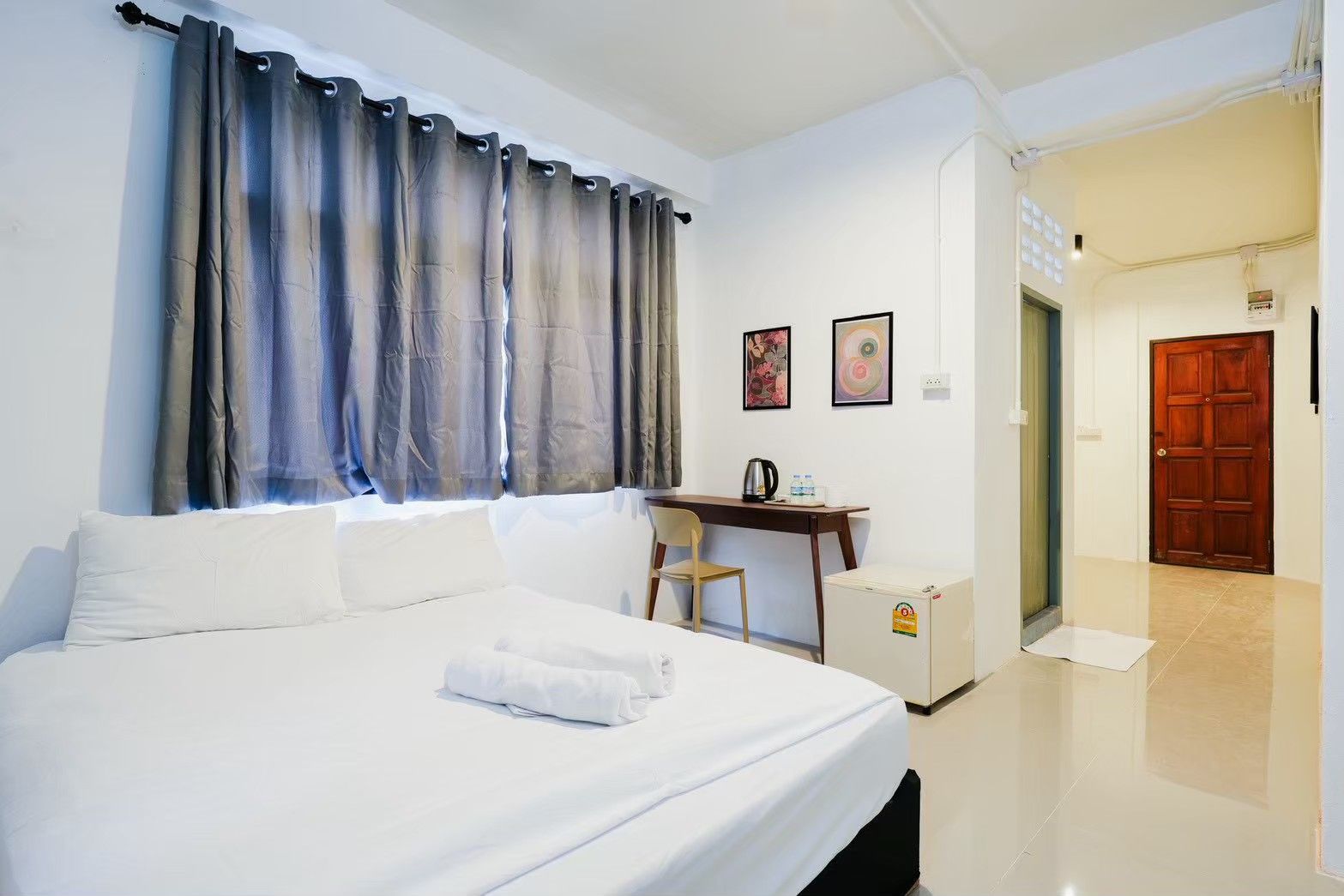A hotel room with a bed, a desk, a chair, a small cabinet, and a door leading to another room.