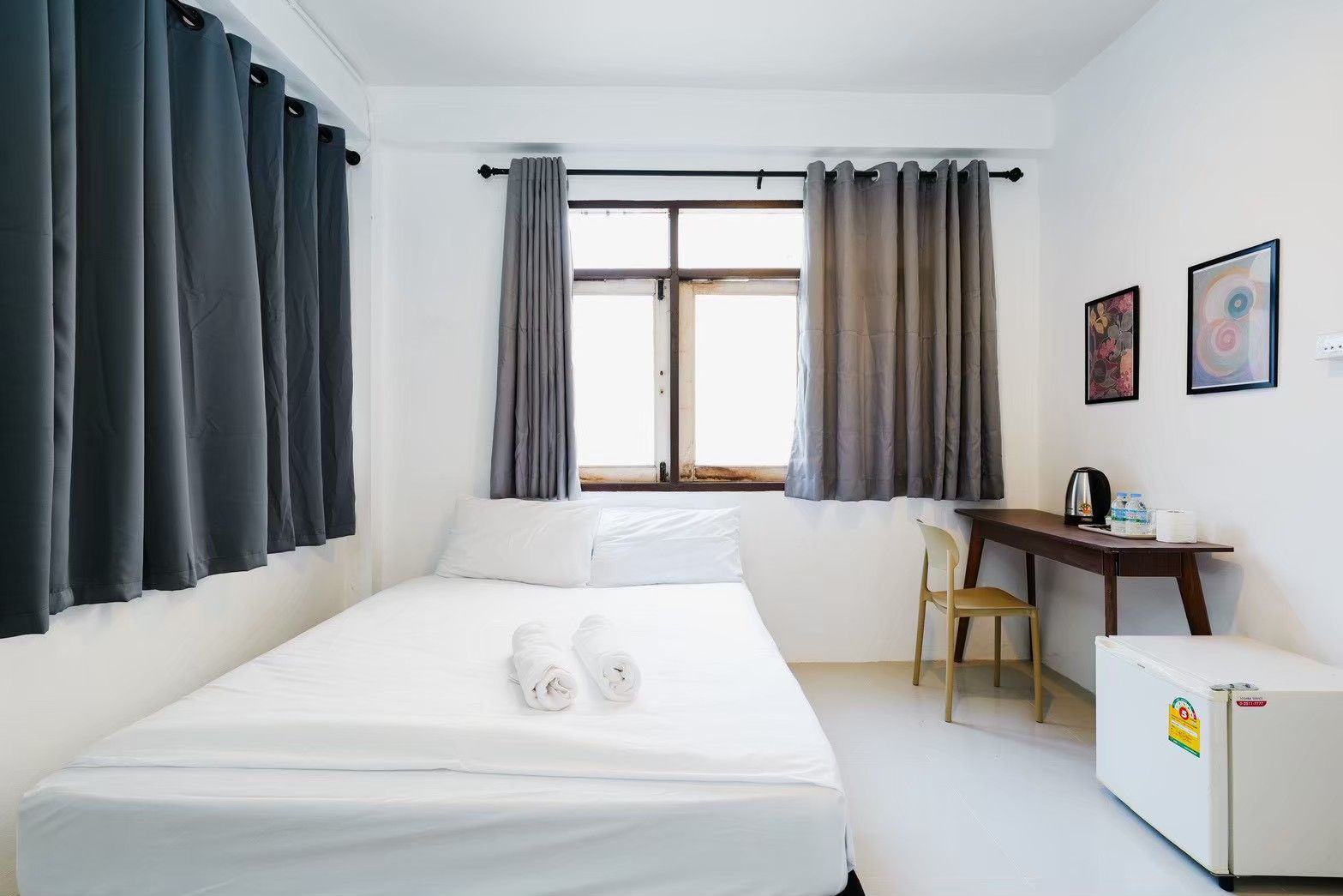 A hotel room with a bed, a desk, a chair, a window with curtains, and a refrigerator.