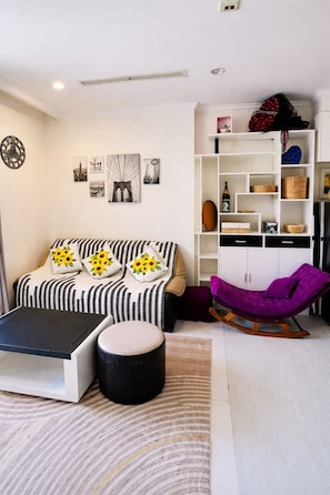 A living room with a sofa, ottoman, and a purple rocking chair. There are wall-mounted shelves with various items and a clock on the wall. - Luxury Apartment Landmark 81 Sakura (Binh Thanh District)