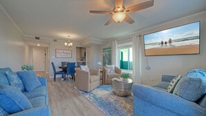 Un salon doté d’un ventilateur de plafond, un coin repas avec des chaises et une table, et une grande fenêtre donnant sur la plage.
