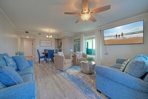 A living room with a ceiling fan, a dining area with chairs and a table, and a large window with a beach view. - Walk to Beach + Boardwalk | Pool + Basketball Access | Sunkissed Seaside (Port Aransas)