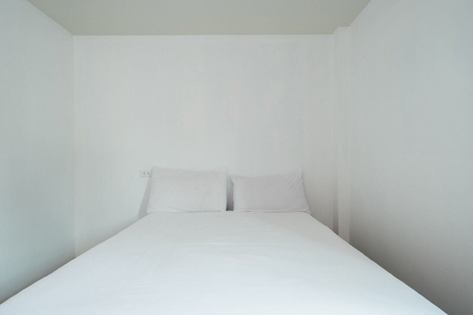 A small, white, single-bed room with a simple, clean design and a visible electrical outlet on the wall.