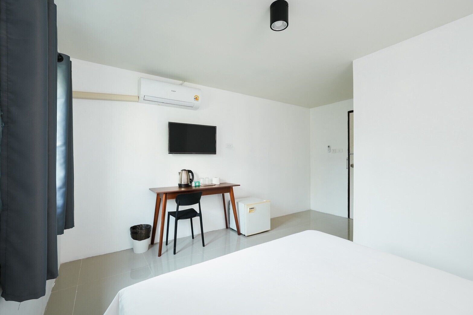 A hotel room with a bed, a desk with a chair, a small refrigerator, a trash bin, an air conditioning unit, and a television on the wall.