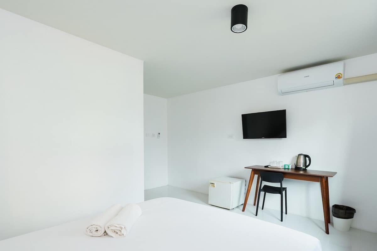 Habitación de hotel minimalista con una cama, un escritorio, una silla, un televisor y un equipo de aire acondicionado.