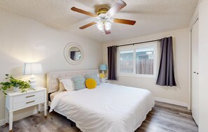 A bedroom with a bed, a nightstand, a lamp, a ceiling fan, a window with curtains, and a wooden floor. - Gorgeous 4-bedroom house with Hot Tub in Huntington Beach I 31 nights + (Huntington Beach)