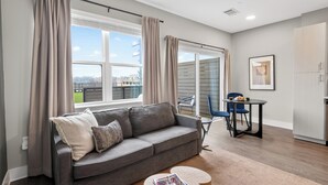 Una sala de estar moderna con un sofá gris, una mesa de centro redonda, una mesa de comedor con sillas y vistas de la ciudad a través de grandes ventanales.