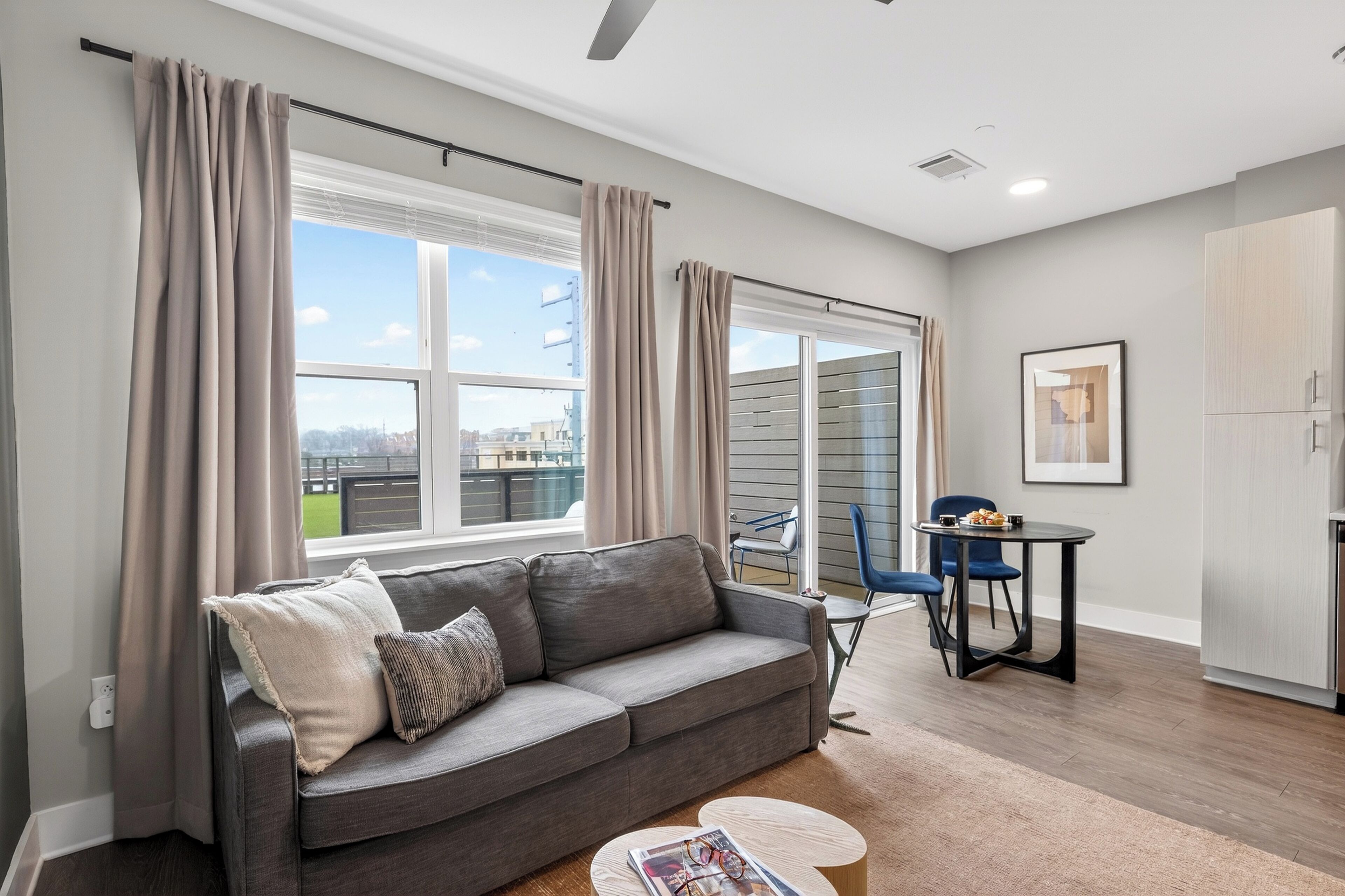 Ein modernes Wohnzimmer mit einem grauen Sofa, einem runden Couchtisch, einem Esstisch mit Stühlen und Blick auf die Stadt durch große Fenster.