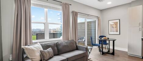 Ein modernes Wohnzimmer mit einem grauen Sofa, einem runden Couchtisch, einem Esstisch mit Stühlen und Blick auf die Stadt durch große Fenster.