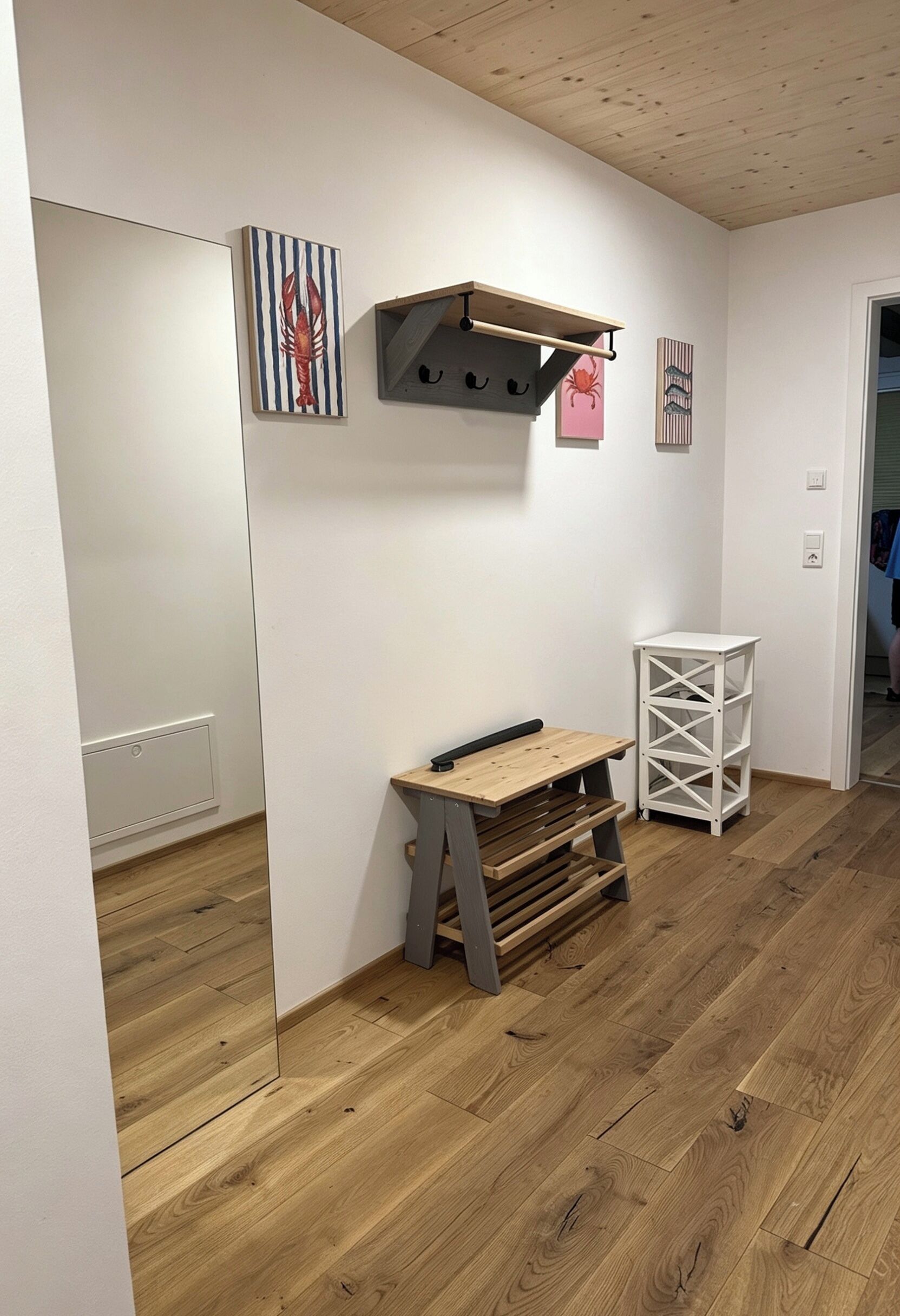 A room with a wooden floor, a white wall, a wooden desk with drawers, a white shelving unit, and a ceiling with exposed beams.