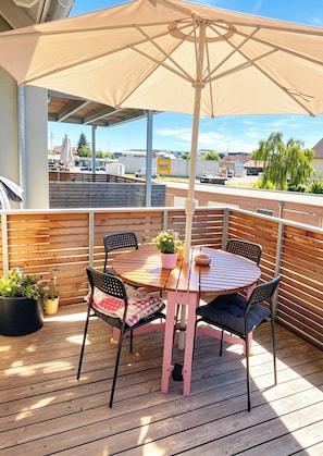 A wooden deck with a round table and chairs, a beige umbrella, and potted plants.
