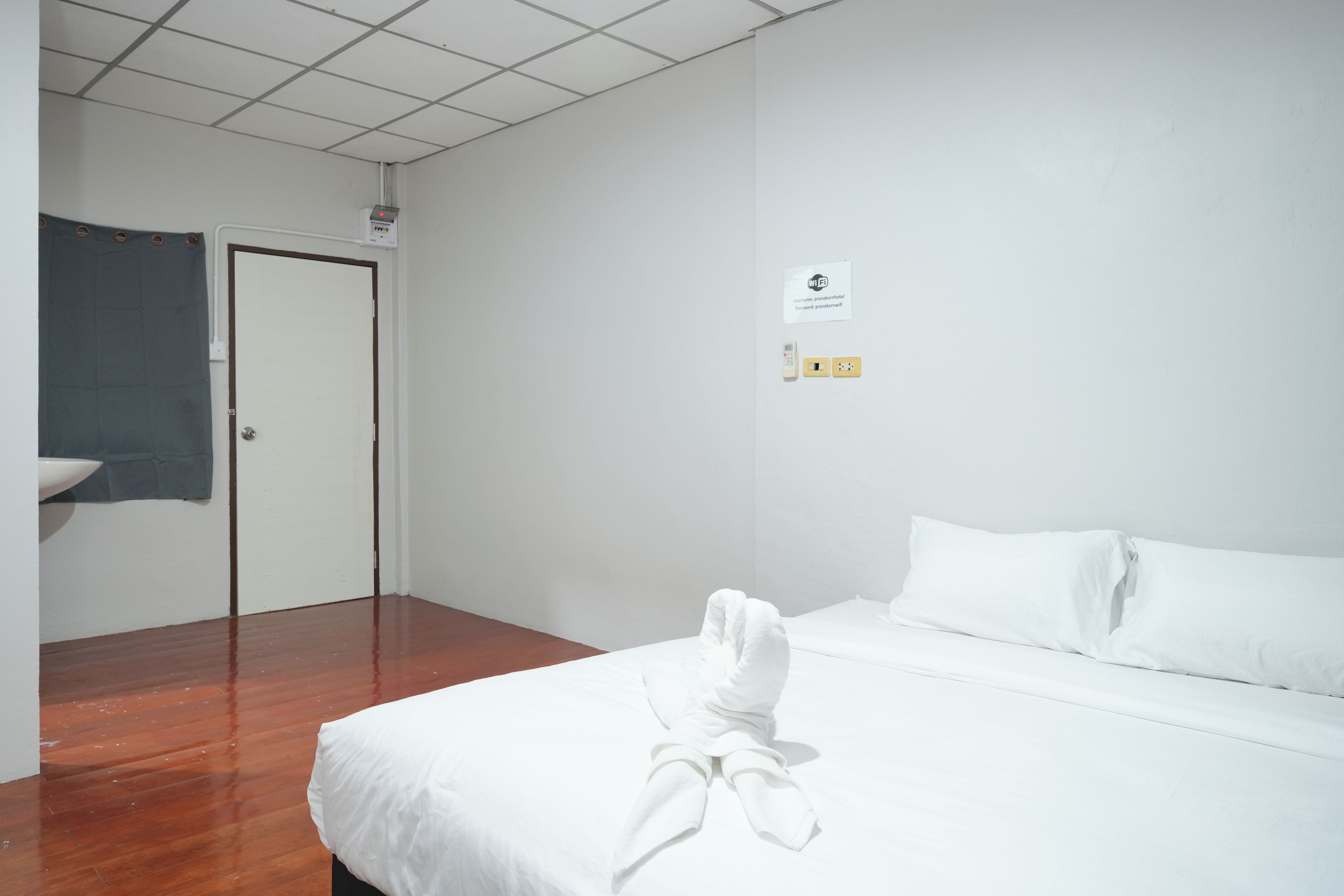 A hotel room with a bed, a door, a towel on the bed, a sign with WiFi details, and a power outlet.