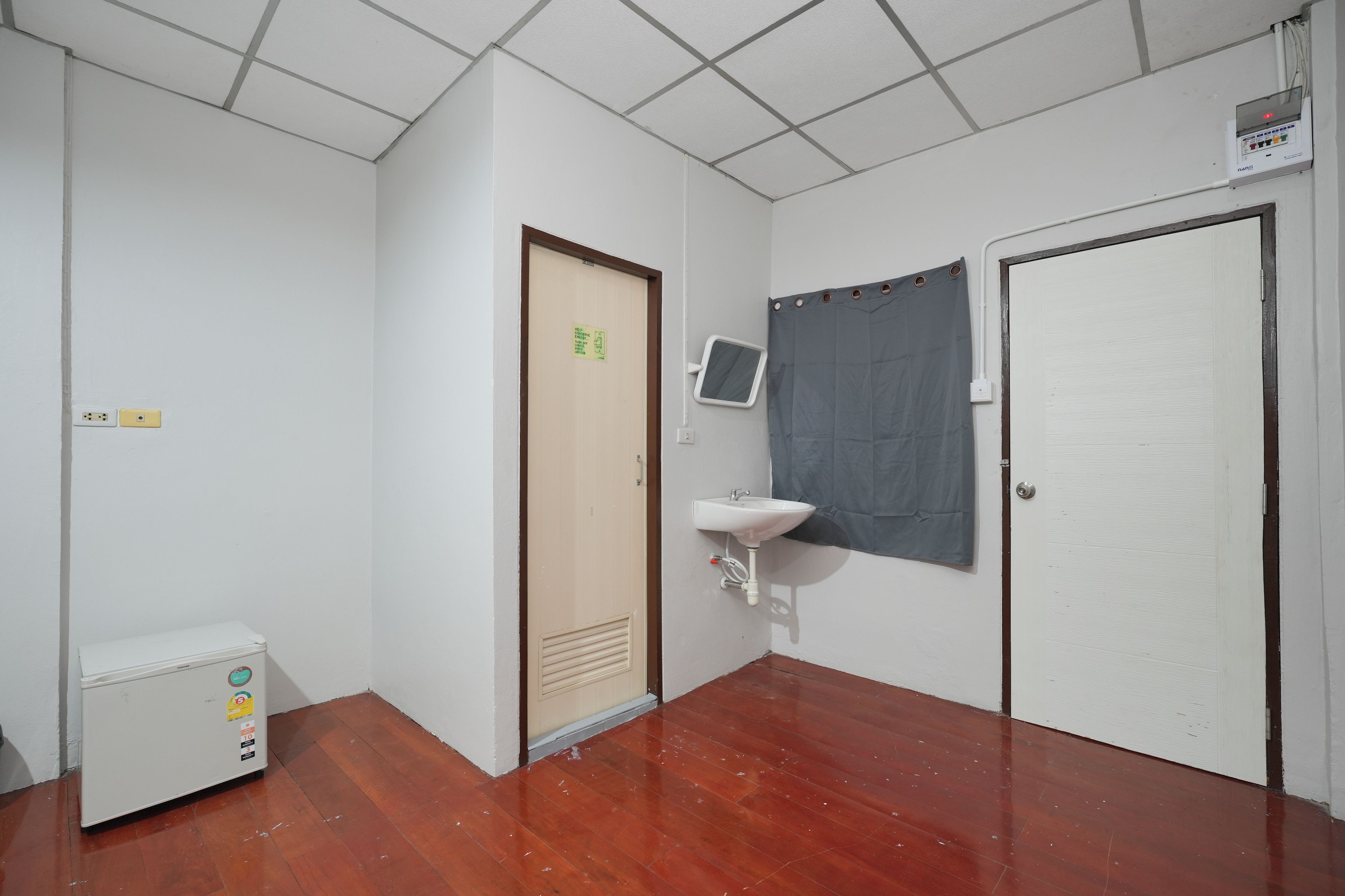 Une petite pièce propre, avec un plancher en bois, un lavabo blanc, une porte dotée d’un miroir et une prise de courant.