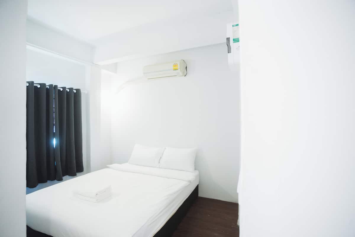 A single bed with white bedding, a dark wooden nightstand, a wall-mounted air conditioner, and black curtains on a white wall.